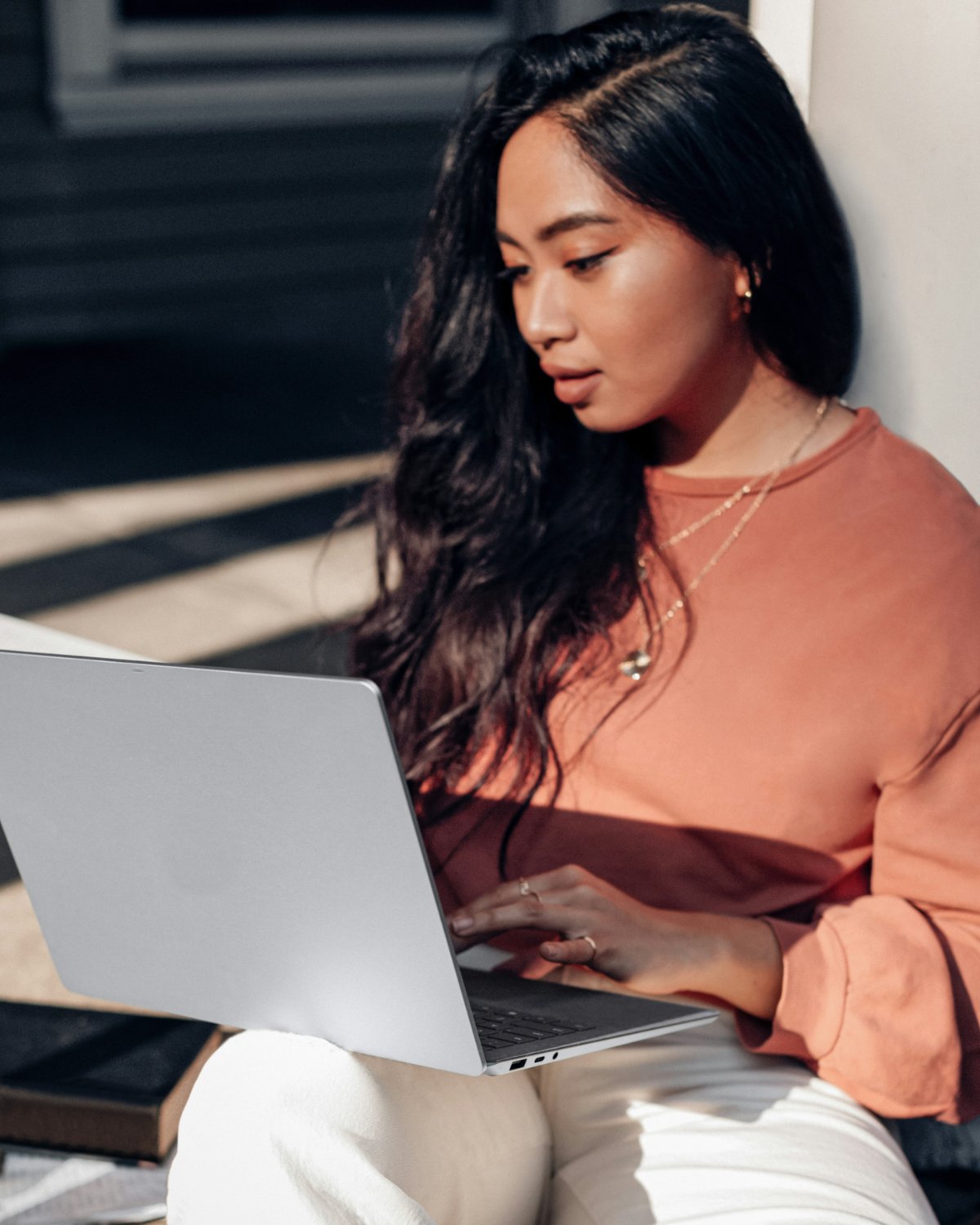 A woman sitting on a sunny doorstep, typing on her laptop